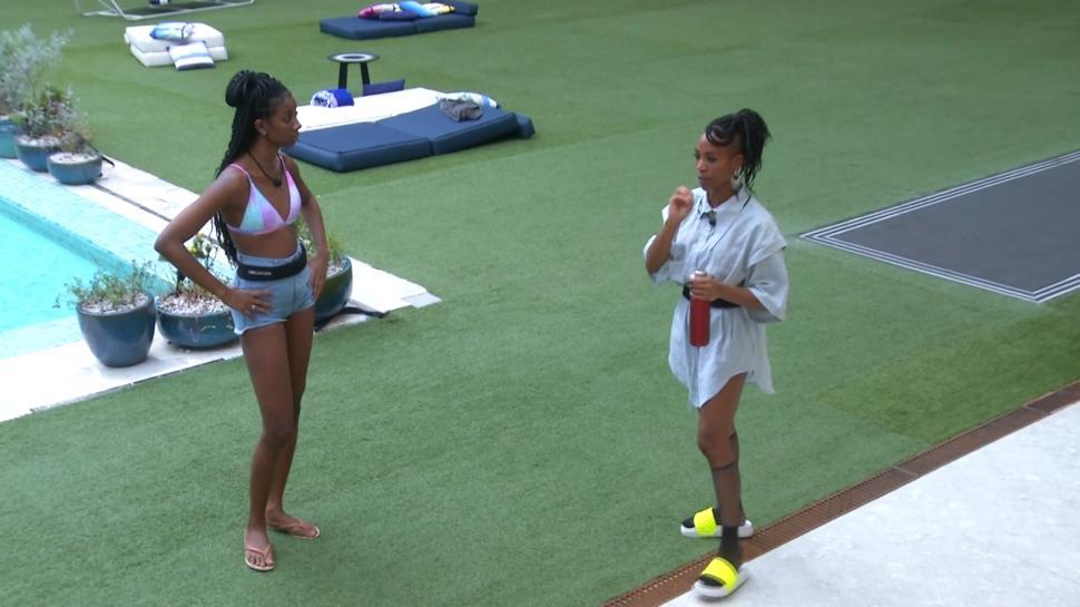 Cena do Big Brother Brasil 21. Camilla e Karol Conká olham uma para a outra no jardim. A câmera mostra as duas de corpo inteiro.