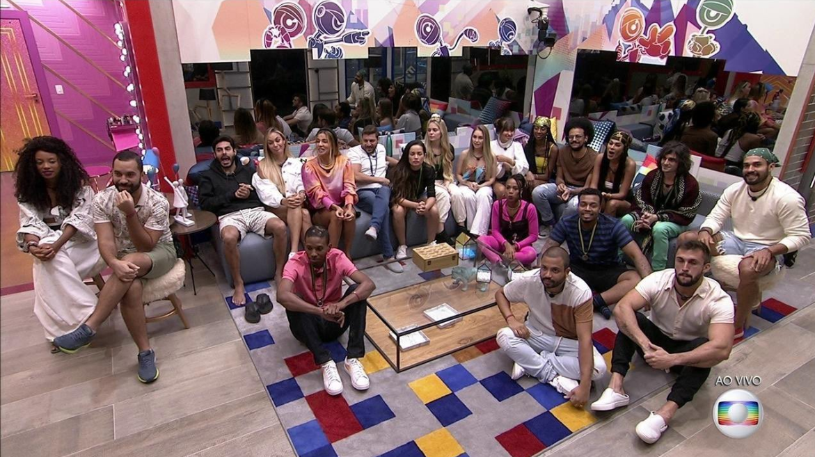Cena do Big Brother Brasil 21. Todos os participantes estão na sala, alguns sentados no sofá e outros sentados no chão. A decoração é colorida, há uma mesa de centro e eles olham em direção ao telão, posicionado embaixo da câmera. 