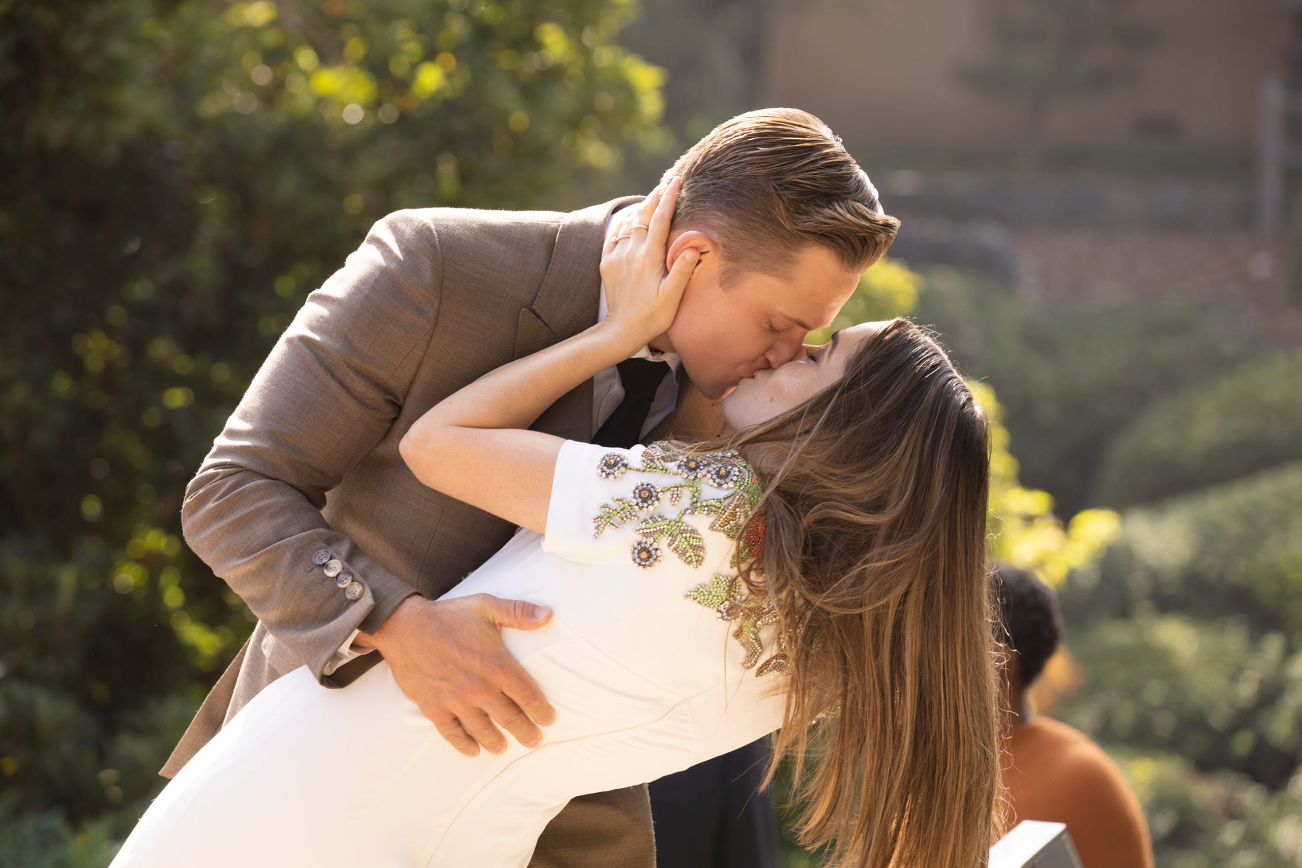 Cena da série Made For Love. No centro da imagem está Cristin Milioti e Billy Magnussen, intérpretes de Hazel Green e Byron Gogol, respectivamente. Cristin é uma mulher branca, de cabelo castanho iluminado longo e solto. Ela usa um vestido branco bordado na parte frontal do busto. Billy é um homem branco de cabelo curto e loiro. Ele veste um terno marrom claro. Na imagem, os dois estão se beijando inclinados no jardim da casa deles.