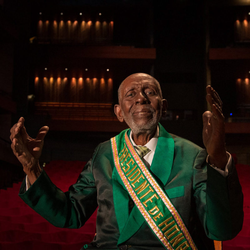 Foto de Nelson Sargento. Nelson é um homem negro de 96 anos, careca com a barba rala branca. Ele está sentado, usa paletó verde e uma faixa escrita PRESIDENTE DE HONRA. Seus braços estão abertos.