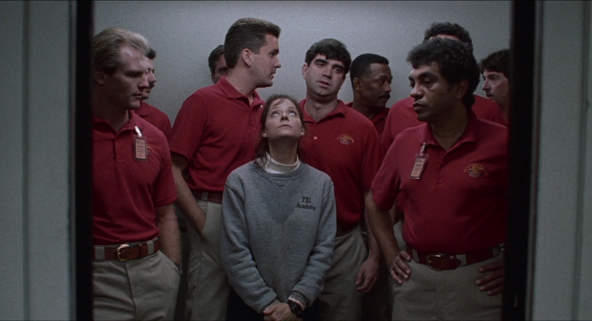 Cena do filme O Silêncio dos Inocentes. Jodie Foster está no centro da imagem, dentro de um elevador. Ela usa moletom cinza com uma camisa branca por baixo marcados com suor, calças pretas e suas mãos estão unidas na frente do corpo. Ao redor dela, vamos nove homens no pequeno espaço. Todos usam a mesma roupa: camisa polo vermelha e calça cáqui bege.
