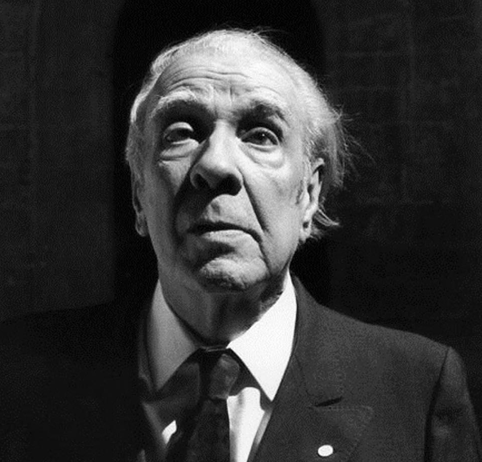 Foto em preto e branco do escritor Jorge Luis Borges. Ele é um homem caucasiano, de cabelos brancos, veste terno preto, camisa branca e gravata preta. Ele mantém a cabeça ereta, enquanto olha para a frente