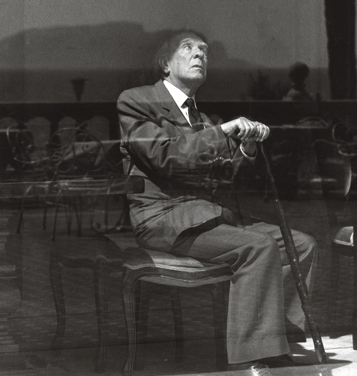 Foto em preto e branco do escritor Jorge Luis Borges. Ele é um homem de cabelos brancos, veste terno e gravata, e está sentado em uma cadeira enquanto segura uma bengala. Está com a cabeça inclinada para trás, olhando para cima