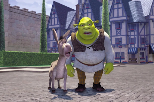 Cena do primeiro filme da franquia de Shrek. A imagem mostra dois personagens principais da trama: Shrek e Burro. Eles estão em uma vila com casas medievais com ornamentos azuis. Ambos estão ao centro da imagem e estão boquiabertos (surpresos). Shrek é um ogro careca, barrigudo e verde. Ele está vestindo camisa de manga longa branca, calça marrom e sapatos pretos. Ao lado esquerdo de Shrek, Burro é um animal quadrúpide cinza que não veste nenhum acessório.