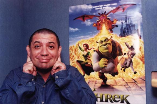 A imagem exibe o ator e comediante Bussunda fazendo um gesto similar às orelhas do ogro Shrek. Bussunda é um homem gordo, branco e careca. Ele veste uma jaqueta jeans azul e está na frente do cartaz oficial do filme que mostra Shrek, Fiona e Burro fugindo do Dragão que cospe fogo.