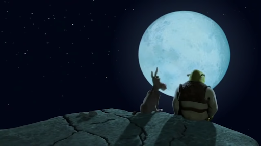 Cena do filme em que Shrek e Burro aparecem sentados na beira de um penhasco refletindo à luz do luar. Ambos estão sentados admirando a lua. Sherek está com sua roupa convencional: camisa longa branca, colete e calças marrons e sapatos pretos. Burro não veste roupa. Shrek está cabisbaixo.