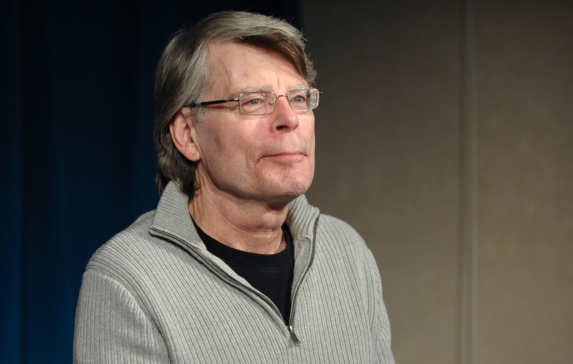 Foto do autor Stephen King. Stephen é um homem branco de 73 anos, com os cabelos levemente grisalhos. Ele usa um moletom cinza com uma camiseta preta por baixo. Possui óculos retangulares. 