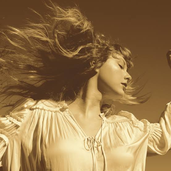 Capa do álbum Fearless (Taylor's Version), de Taylor Swift. A capa é composta unicamente por uma imagem de Taylor, uma mulher branca e loira, que aparece sobre o centro de um fundo da amarronzado, sépia. Aparentemente saltando com o cabelo ao vento.Aqui ela usa uma blusa branca
