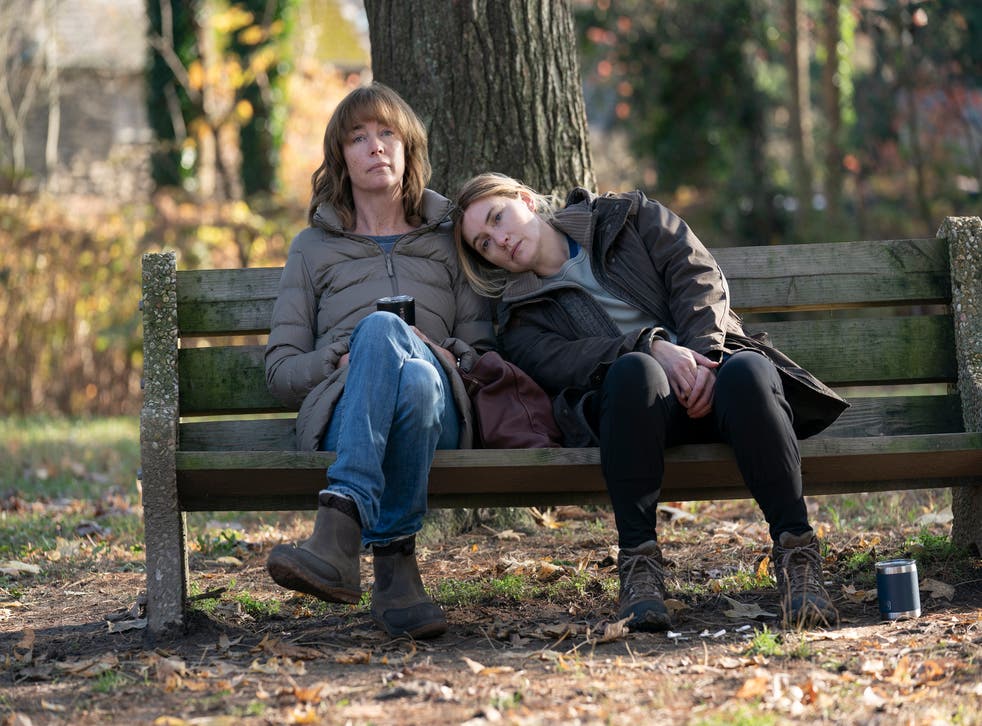 Cena da série Mare of Easttown. Na cena, vemos Lori e Mare sentadas em um banco de praça, de dia, e ao fundo está cheio de árvores e mato. As duas são brancas, tem cabelos escuros e usam roupa de frio. 