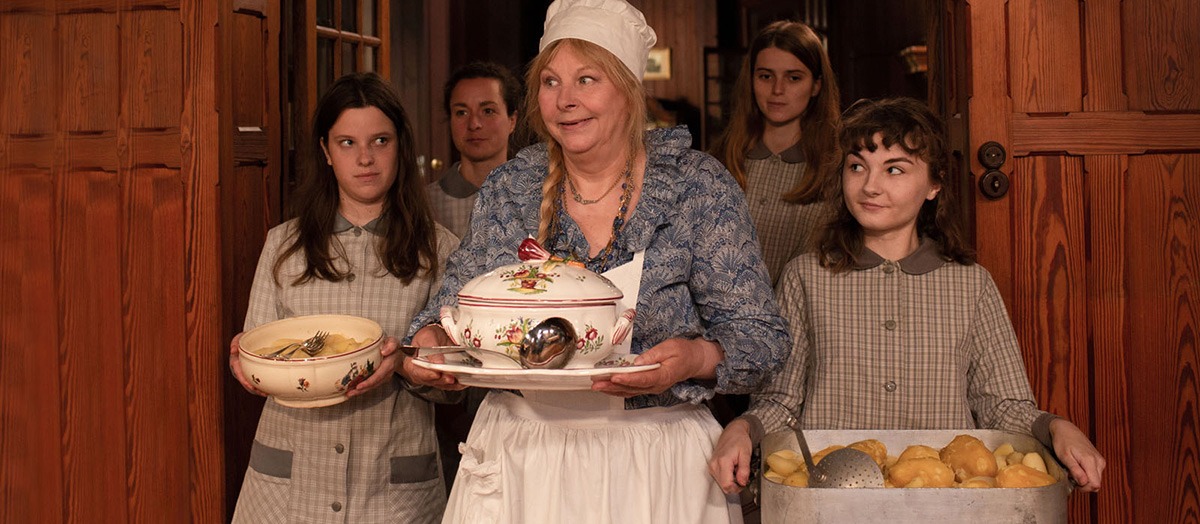 Cena do filme A Boa Esposa. Uma mulher mais velha está no centro da imagem, com roupas de cozinheira segurando um prato de porcelana. Ao seu redor, garotas jovens uniformizadas a olham com orgulho.