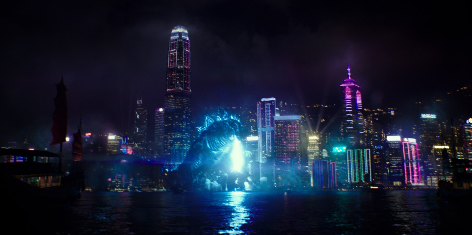 Cena do filme Godzilla vs Kong. A imagem é retangular, e no centro dela está Godzilla, que brilha em azul, seu rosto está virado para baixo e sua boca está aberta, de dentro dela sai um raio azul. Ele está no meio de uma cidade e ao seu redor há vários prédios que brilham em cores vibrantes. O fundo da imagem é o céu noturno. 