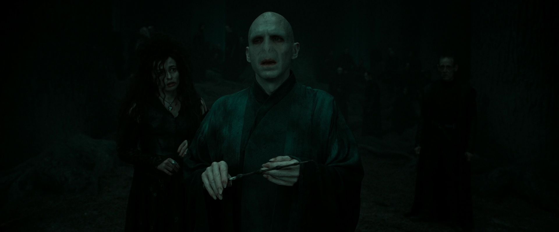Cena do filme Harry Potter e as Relíquias da Morte: Parte 2. A cena mostra Voldemort na floresta, no escuro e com a Varinha das Varinhas na mão. Atrás dele, e meio desfocada, está Bellatrix Lestrange. Voldemort é careca, tem pele cor de leite e um nariz de cobra. Bellatrix é branca, tem cabelos sedosos pretos e usa um vestido escuro.