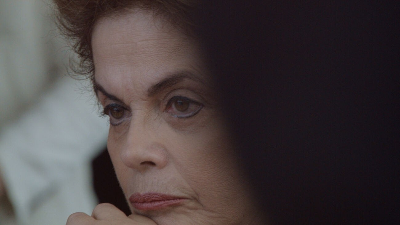 Cena do filme Alvorada. A imagem é retangular e mostra o lado esquerdo do rosto de Dilma Rousseff. Ela é uma mulher branca, de olhos castanhos e cabelos também castanhos curtos. Dilma usa maquiagem preta no contorno dos olhos e um batom rosa cor de boca. Dilma está olhando para fora da imagem, para o lado esquerdo, com o rosto inclinado. Na diagonal direita, da metade da foto em diante, existe uma sombra escura.