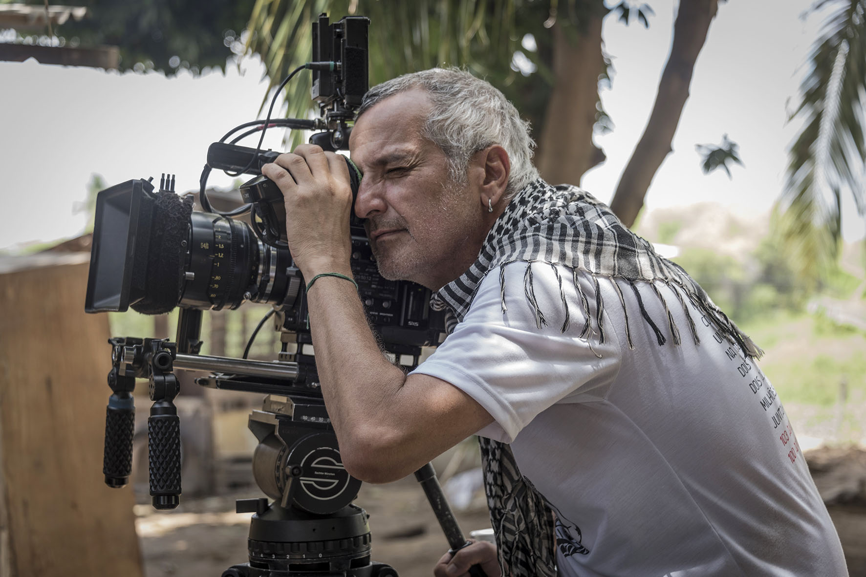 Foto do diretor Lírio Ferreira. Ele está de perfil, olhando pela lente de uma câmera de filmagem em um tripé. Lírio é um homem branco de 50 anos, com cabelos grisalhos e ralos. Ele usa uma camiseta branca e um chale por cima.