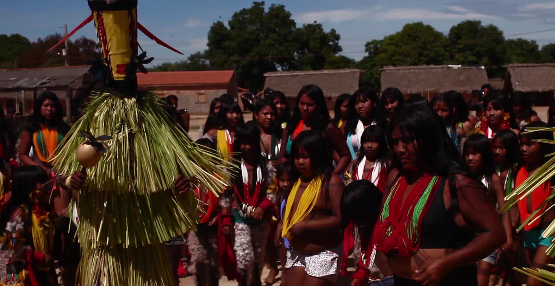 Cena do filme Apyãwa (Tapirapé) Iraxao Rarywa que exibe diversos indígenas, em sua maioria mulheres, vestindo roupas típicas de sua cultura, em volta de uma pessoa que usa uma vestimenta desfiada e toca um chocalho. O fundo é composto por casas, árvores e o céu azul.