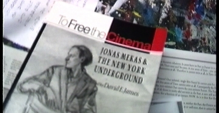 Cena do filme Com Amor: Volume 1 1987-1996 exibe livros espalhados, sendo que um deles se chama Para Libertar o Cinema, em inglês, e apresenta o desenho de um homem sentado em uma cadeira olhando para trás.