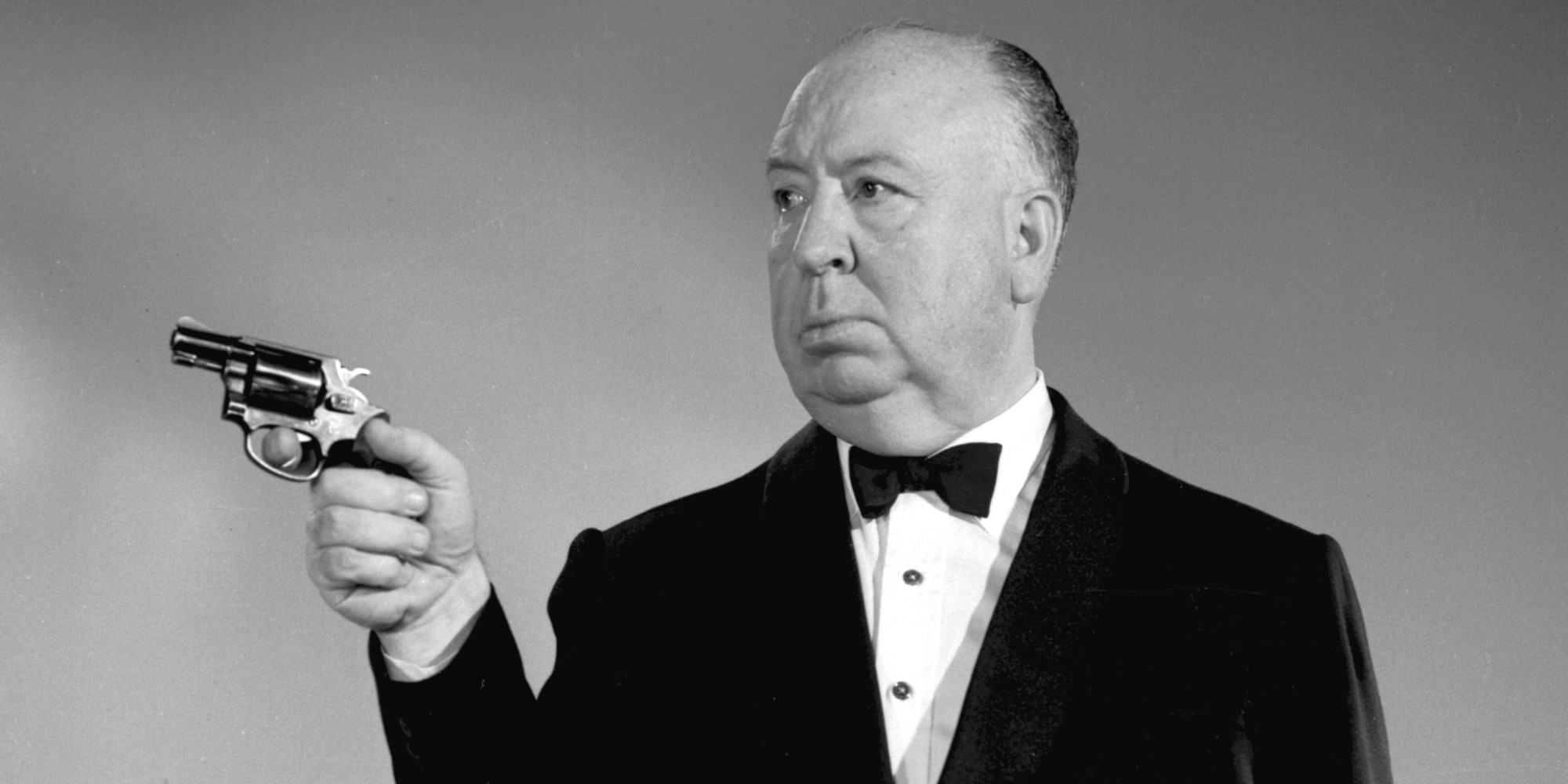 Vemos uma foto em preto e branco do Diretor Alfred Hitchcock, falecido em 1980. Ele é um homem branco, calvo e robusto que usa um smoking preto e aponta uma pistola para a nossa esquerda