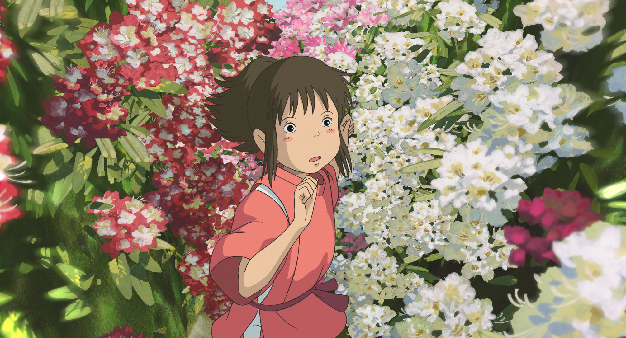 Imagem do filme de animação “A Viagem de Chihiro”. A cena se passa de dia. No centro, vemos Chihiro, uma garota asiática, de pele branca, bochechas rosadas e cabelos castanhos presos em um rabo de cavalo, vestindo um kimono vermelho. Ela se esgueira entre um grande jardim de flores brancas e vermelhas, e seu semblante é confuso.