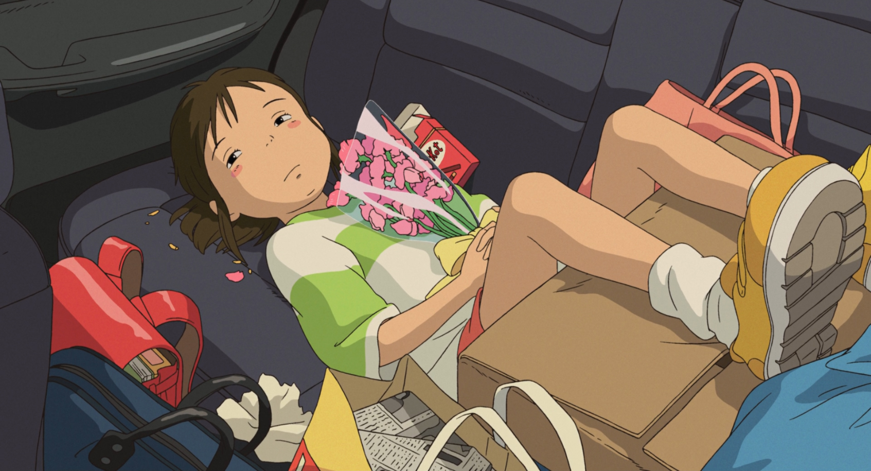Imagem do filme de animação “A Viagem de Chihiro”. A cena se passa de dia. Ocupando quase toda a imagem, está Chihiro, uma garota asiática, de pele branca, bochechas rosadas e cabelos castanhos presos em um rabo de cavalo. Ela veste uma camisa branca e verde, um short vermelho e tênis amarelos. Ela está deitada no banco de trás de um carro, que está abarrotado de malas e sacolas, com os pés apoiados em uma grande caixa de papelão. A menina olha para o lado, triste e melancólica, enquanto segura com as duas mãos um buquê de flores rosadas, pressionando-as contra seu peito.