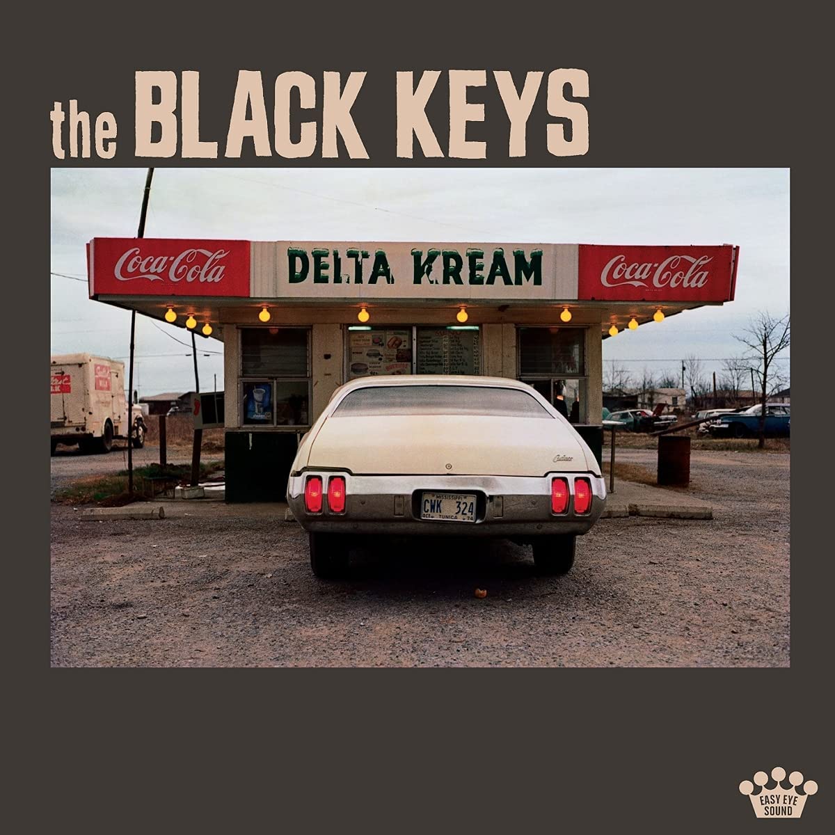 Capa do CD Delta Kream. Apresenta o título da banda The Black Keys acima. Em uma fotografia quadrada, há um carro branco estacionado no centro, em frente a uma loja chamada Delta Kream, que possui dois símbolos da Coca-Cola ao lado do nome. O chão é de terra batida.
