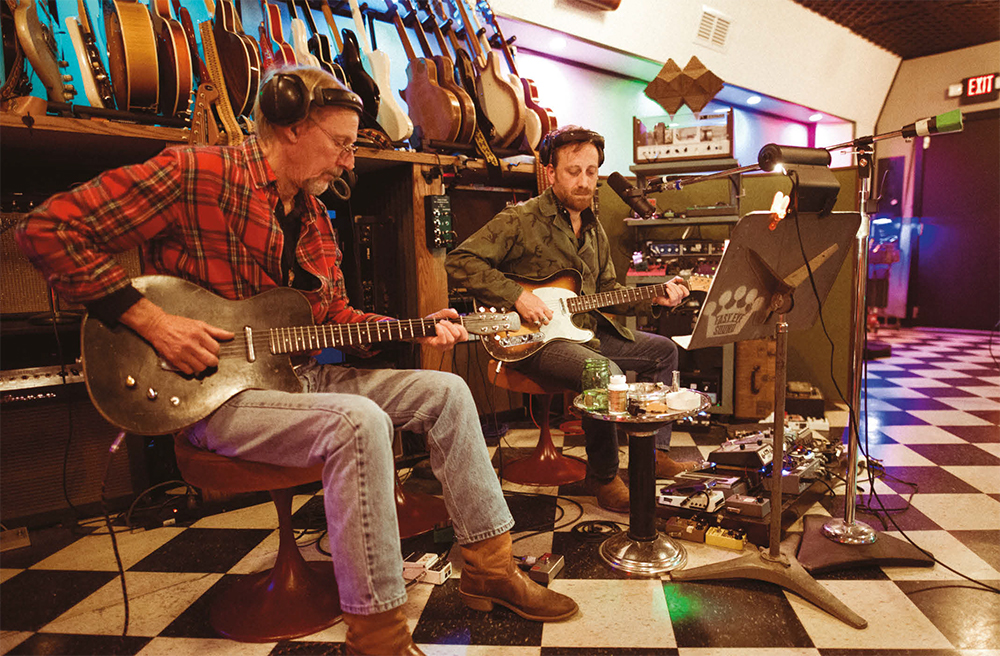 Foto de Kenny Brown e Dan Auerbach. Os dois são homens brancos e estão em um estúdio, com guitarras penduradas ao fundo. Kenny é mais velho que Dan, está com fones de ouvido na cor preta, camisa xadrez vermelha, branca e preta, e toca uma guitarra preta. Está usando uma calça jeans clara e botas na cor marrom. Dan está com fones de ouvido na cor preta, uma camisa verde, tocando uma guitarra branca, laranja e preta, com uma maleta de pedais aberta no chão. Ele usa uma calça jeans escura e botas na cor marrom. Ambos estão sentados em bancos da cor marrom.