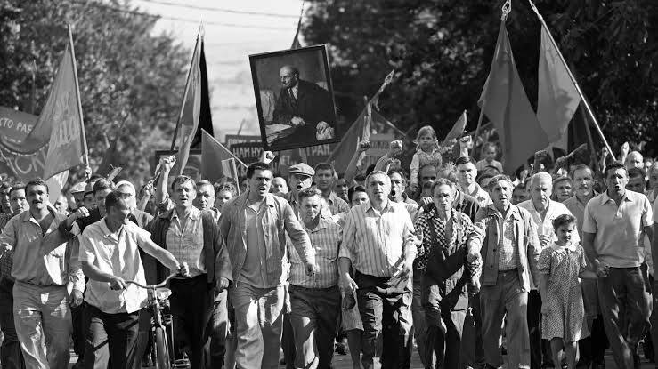 cena do filme Caros Camaradas! em branco e preto na qual os trabalhadores vão às ruas para protestar. A imagem apresenta dezenas de proletários, majoritariamente homens, erguendo bandeiras e um quadro de Lenin.