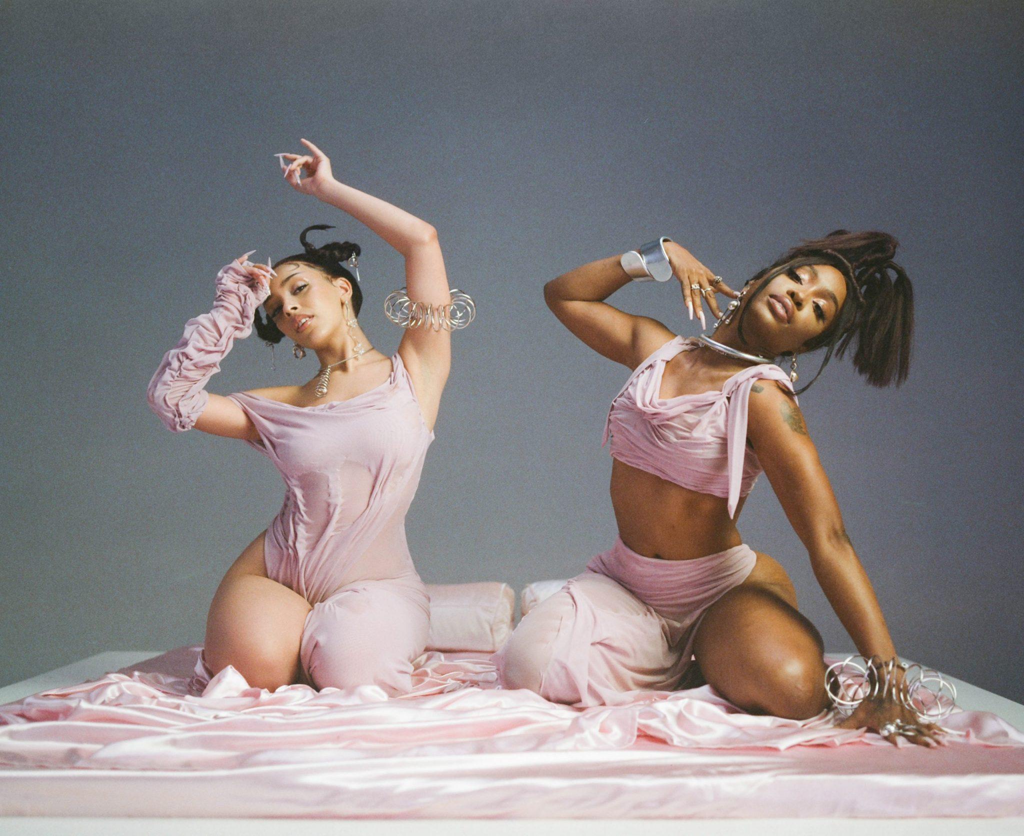 Fotografia da produção do clipe da música Kiss Me More, da cantora Doja Cat em parceria com SZA. A foto mostra as cantoras, duas mulheres negras de cabelos e olhos castanhos, vestindo roupas cor de rosa, sentadas em uma cama.