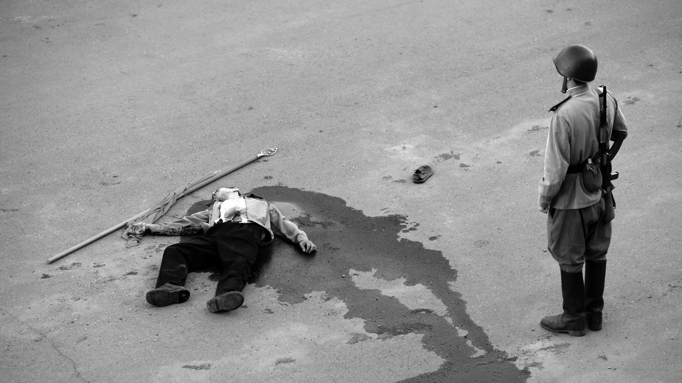 cena do filme Caros Camaradas! em branco e preto que mostra um homem morto baleado no chão, com o sangue escorrendo ao seu redor e uma bandeira perto de sua mão. No canto direito está um soldado de costas para a imagem vestindo a farda do exército e olhando para o homem baleado no chão.
