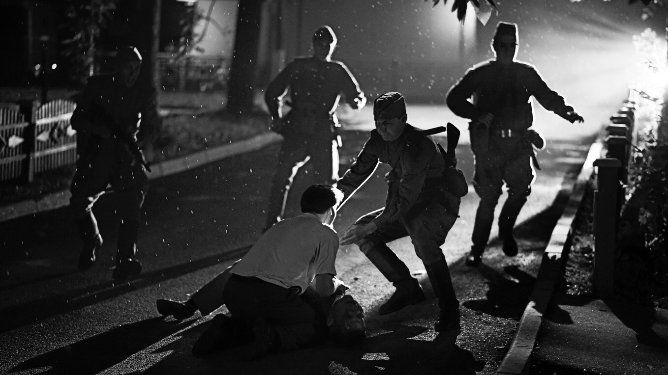 cena do filme Caros Camaradas! em branco e preto em que um homem tenta salvar outro ferido. Ao redor estão quatro soldados, que correm em direção aos protestante no chão.