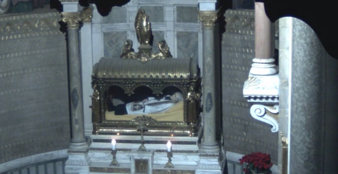 Cena do filme Natalis apresenta uma igreja com um santo dentro de uma redoma de vidro e metal, usando uma batina branca e uma estola vermelha. Na frente da cúpula está uma cruz pequena de metal, duas velas e duas colunas na lateral.
