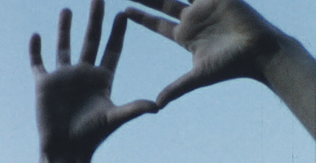 Cena do filme O Céu Socialista que exibe duas mãos de uma pessoa branca unindo os indicadores e o polar para formar um triângulo. Ao fundo é possível ver o céu azul.