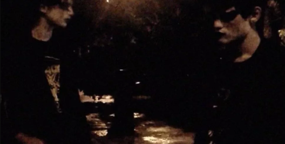 Cena do filme Sombra exibe dois rapazes vestidos somente com roupas pretas. Usam maquiagem branca e preta ao redor dos olhos. Um está de frente para o outro em uma praça, durante a noite.