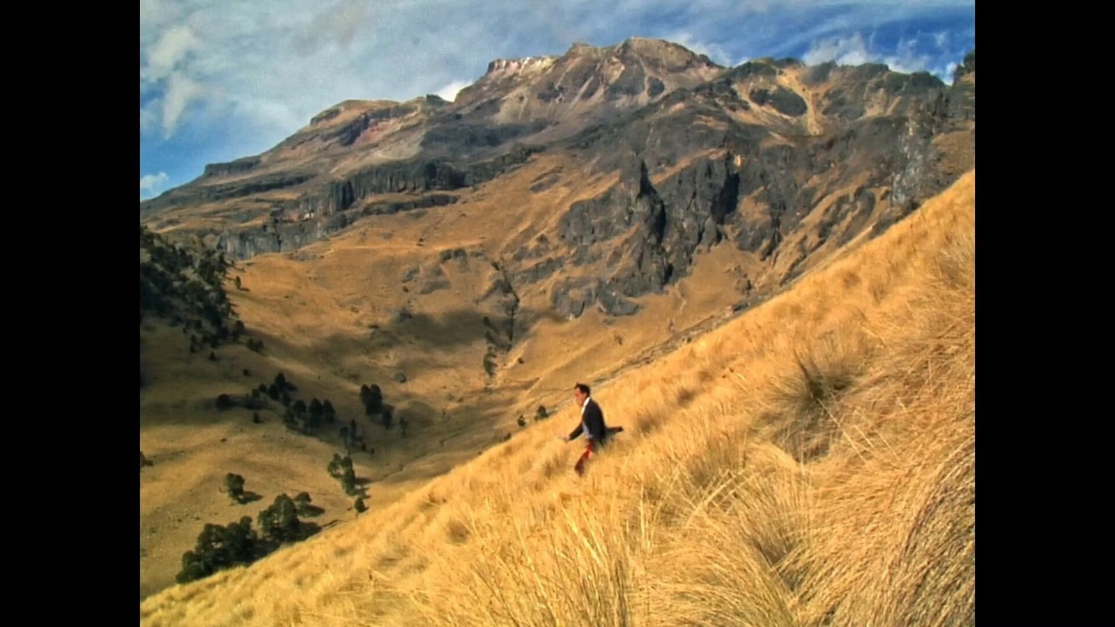 Cena do filme Toda Luz Que Podemos Ver exibe uma paisagem com vegetação rasteira e seca, montanhas ao fundo e o céu azul com poucas nuvens. Ao centro da imagem aparece um homem de roupa preta andando pela vegetação baixa.