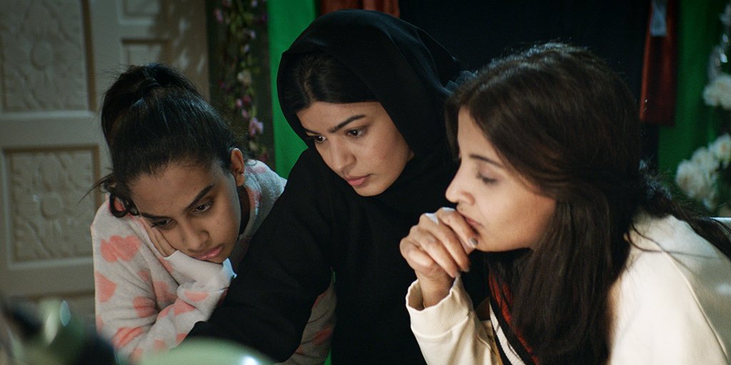Cena de A Candidata Perfeita. Nele estão três as mulheres protagonistas olhando para a tela de um computador com as expressões sérias. A do meio veste roupa e hijab preto enquanto as dos lados estão de roupas rosas claras.