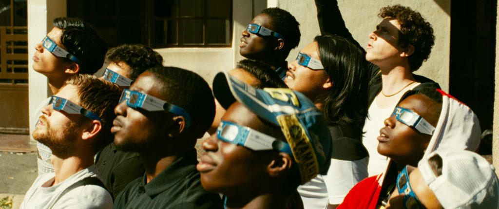 Cena do filme Edifício Gagarine. A imagem mostra um grupo de jovens de diferentes etnias, todos voltados para o lado esquerdo, olhando para cima. Eles usam óculos iguais, com as cores azul, branco e preto. 