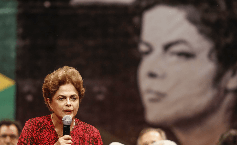 Cena do filme O Processo. A imagem mostra Dilma Rousseff no canto inferior esquerdo. Ela veste uma blusa vermelha e segura um microfone com a mão esquerda, enquanto olha para a frente e fala. Ao fundo, desfocada, existe uma imagem dela enquanto mais nova em preto e branco.