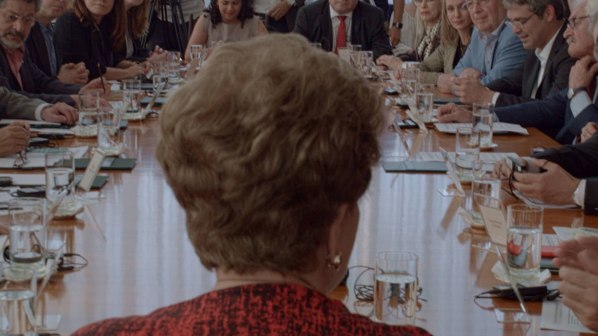 Cena do filme Alvorada. A imagem mostra a presidenta Dilma Rousseff de costas, numa mesa de reuniões repleta de pessoas. Dilma usa um blazer vermelho com desenhos pretos e olha para a frente, onde está a mesa. Todos olham para ela ao redor da mesa, onde existem vários copos de água, papéis e dispositivos eletrônicos.
