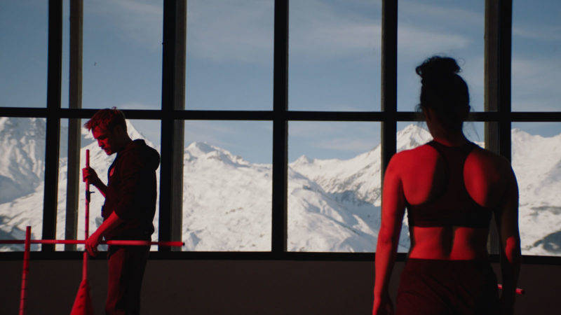 Cena do filme Slalom. À esquerda está o treinador. Um homem branco e de cabelos ruivos. À direita está Lyz de costas. O fundo é uma janela que mostra a paisagem de montanhas. Há uma luz vermelha em toda a cena.