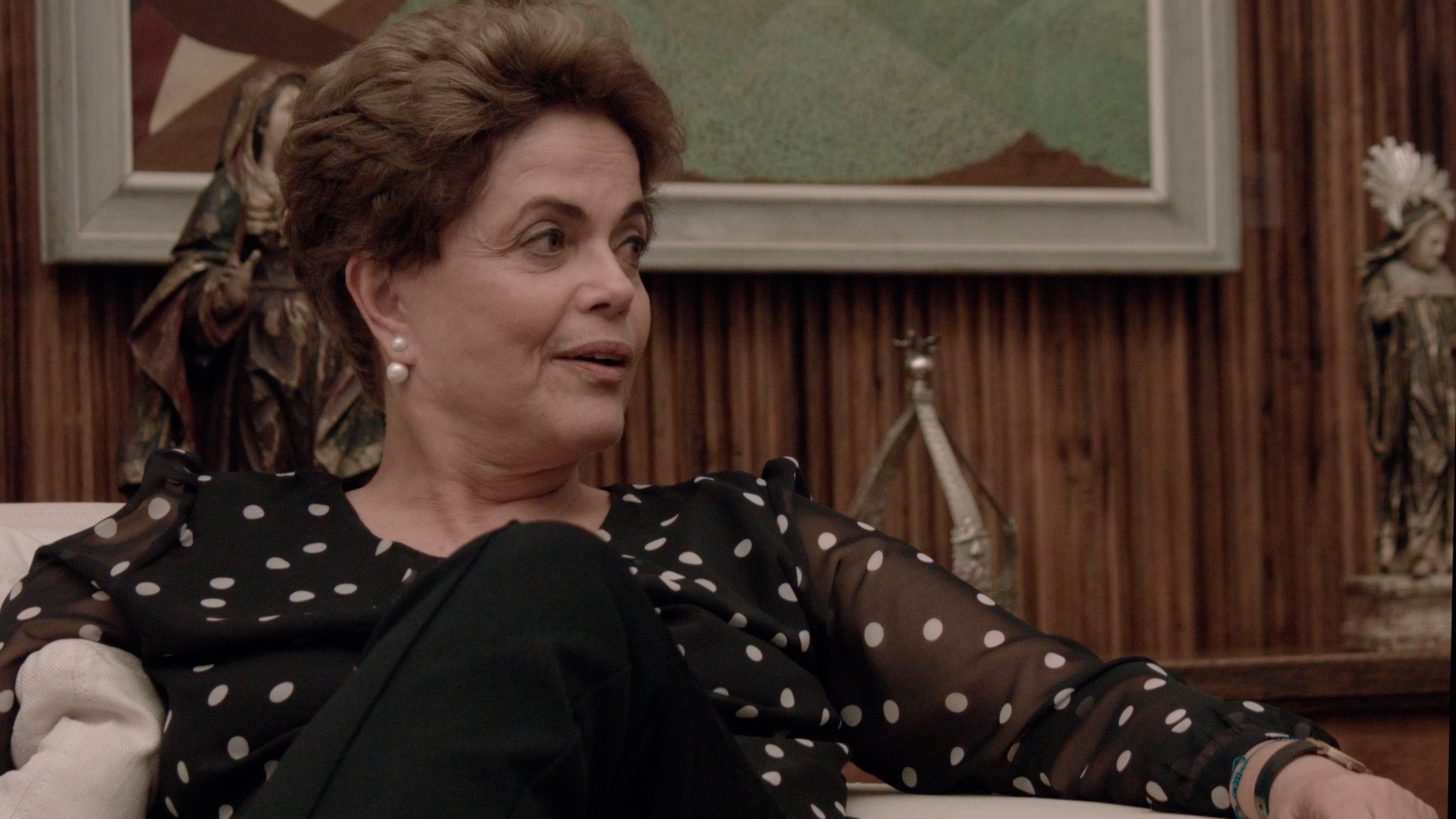 Cena do filme Alvorada. A imagem mostra a ex-presidenta Dilma Rouseff sentada num sofá, com as pernas cruzadas, conversando. Dilma, uma mulher branca, de cabelos curtos loiros escuros, usa uma camisa preta de bolinhas brancas e calça preta, e um brinco de pérola. Dilma olha para o lado direito da imagem, e ao fundo, existe um quadro e uma estante com enfeites de metal.