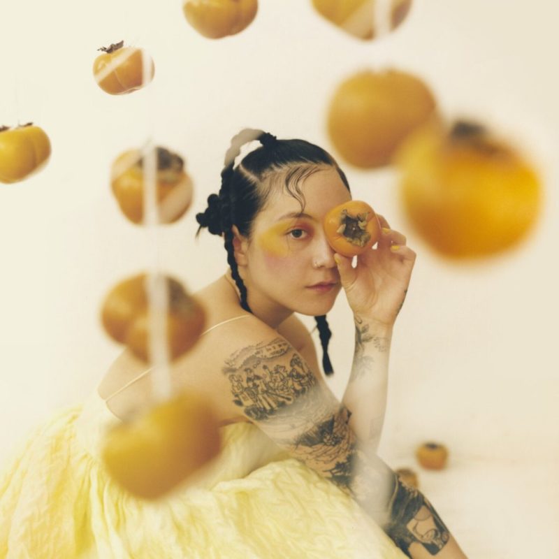 A cantora Michelle Zauner na capa do disco Jubilee vestindo um vestido amarelo, maquiagem artística no mesmo tom e tranças no cabelo. Há caquis pendurados no primeiro plano e ela segura um na frente do olho direito.O fundo é claro, quase branco. Há também muitas tatuagens em seus braços.