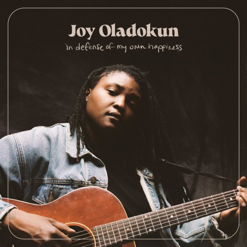 Capa do CD In Defense of My Own Happiness, da cantora Joy Oladokun, uma mulher negra, que está na foto de jaqueta jeans e tocando violão