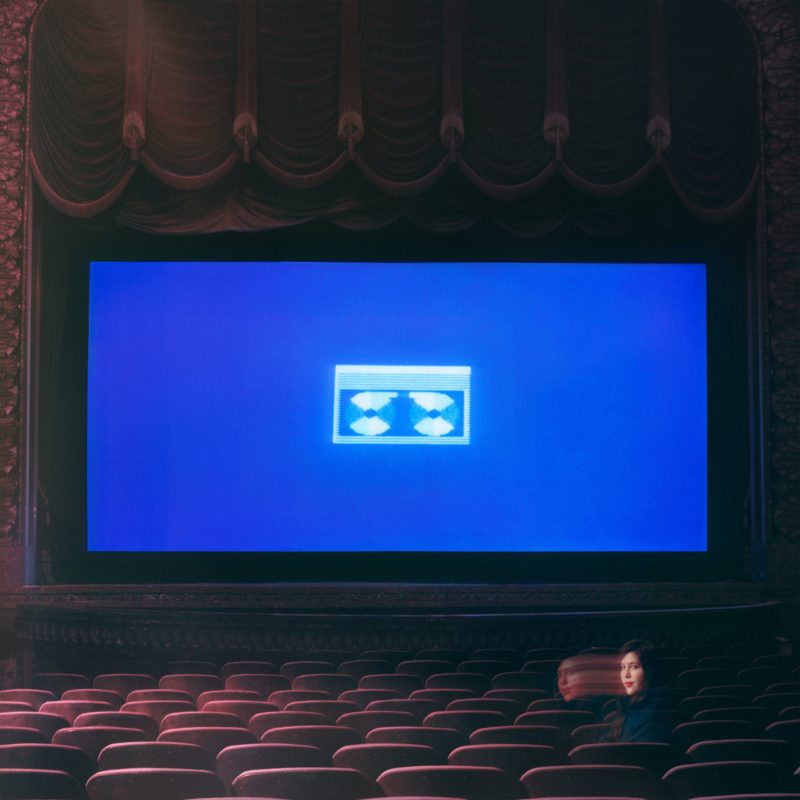 Capa do CD Home Video, da cantora Lucy Dacus. A imagem mostra uma tela de cinema, com um desenho de fita cassete em fundo azul. Nos bancos do cinema, está Lucy, com o rosto sombreado e enigmático. 