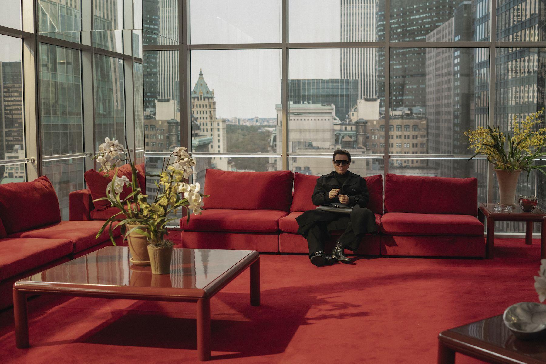 Cena da série Halston. Ewan McGregor está no fundo da imagem, vestido inteiramente de preto. Ele está sentado em um sofá vermelo, que combina com o carpete também vermelho da sala. Ao seu redor, todas as paredes são de vidro, assim como a mesinha no canto esquerdo, que contém um vaso com orquídeas.