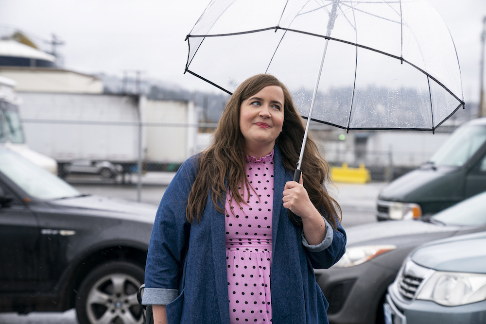 Cena da série Shrill. Na cena, vemos Aidy Bryant, uma mulher branca e gorda, parada na rua segurando um guarda-chuva de plástico transparente. Ela está com uma expressão feliz, usa uma blusa rosa e um casaco azul. Está de dia.
