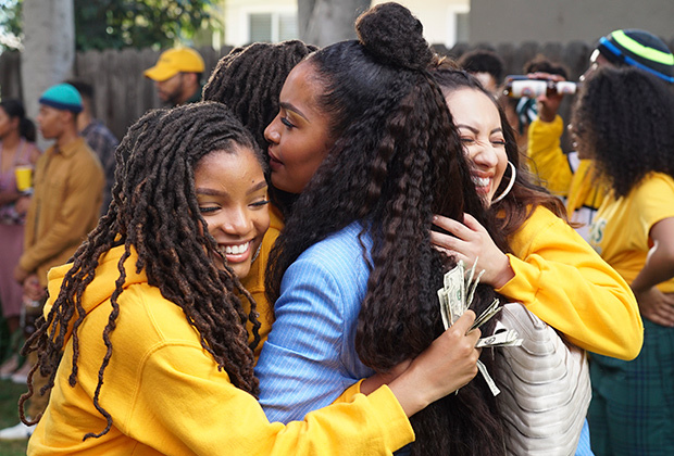 Cena de grown-ish. Nela está uma menina negra, de cabelos cacheados longos com uma parte presa em um coque, sendo abraçada por outras três meninas, duas negras e uma branca, todas usando um moletom amarelo. As meninas sorriem. No fundo é possível ver pessoas espalhadas por um jardim.