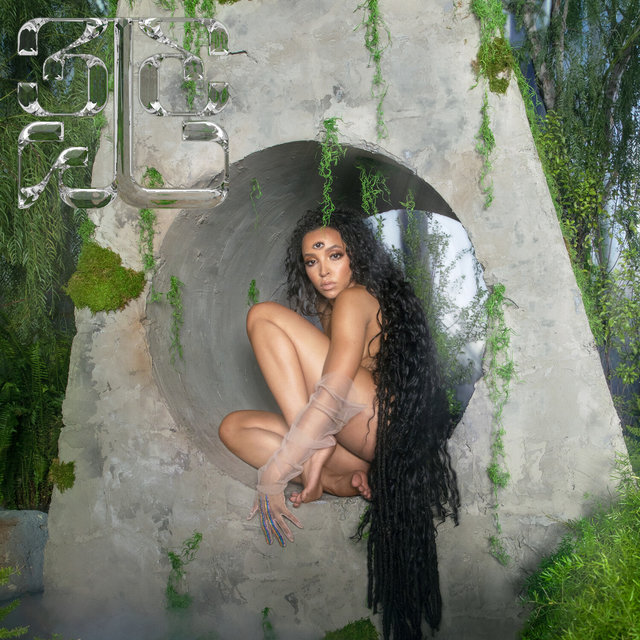 Capa do álbum 333 da cantora Tinashe. Em uma floresta esverdeada, Tinashe se senta sobre um bloco de pedra triangular que contém um círculo em seu meio. Ela se posiciona com as pernas dobradas para o lado direito. Tinashe é uma mulher negra, e seus cabelos são escuros e longos. Em sua testa, um terceiro olho se revela sobre sua pele.