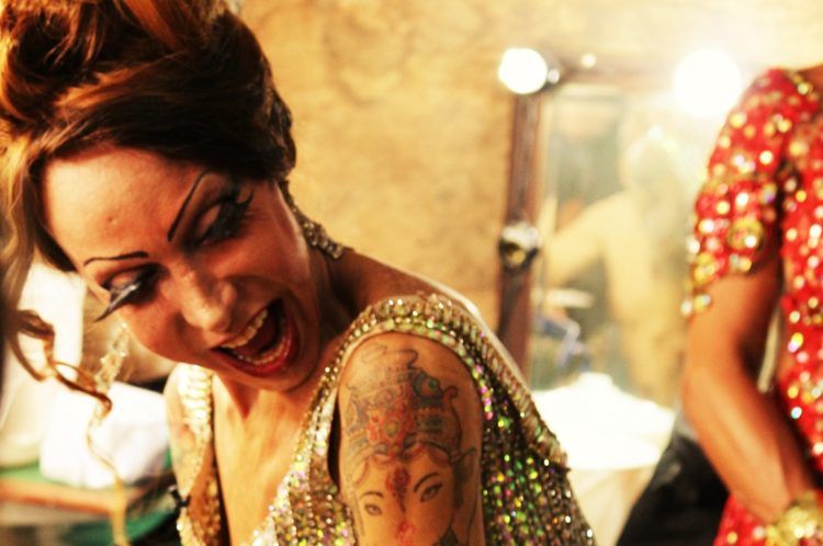 Cena do filme Luana Muniz - Filha da Lua. A foto mostra Luana dentro do camarim, rindo. Vemos sua boca aberta, feliz, e uma tatuagem no braço direito. Ao seu lado, alguém está cortado da imagem. 