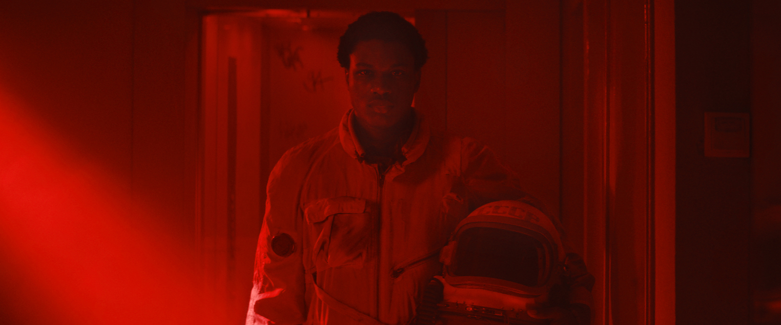 Cena do filme Edifício Gagarine. O personagem Youri, um menino de 16 anos negro de pele escura, está vestido com uma roupa de astronauta vintage. Ele segura o capacete da roupa nos braços. Ele está em um corredor, caminhando em direção a câmera. A iluminação da cena é toda em vermelho. 