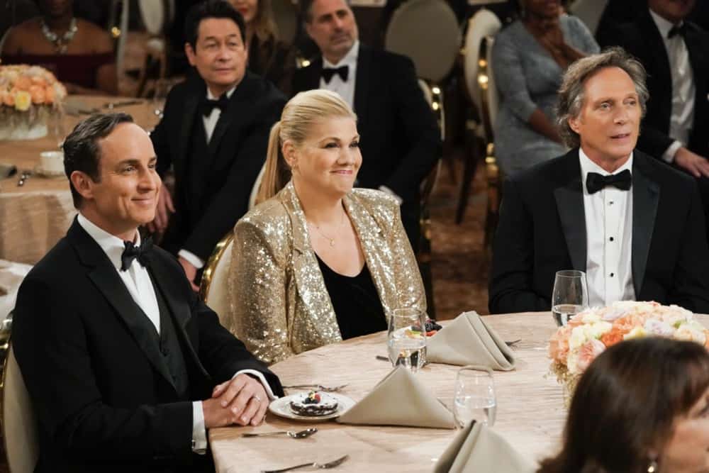 Cena da série Mom, que mostra 3 pessoas brancas sentada à uma mesa chique, usando roupas de gala e olhando para um palco.
