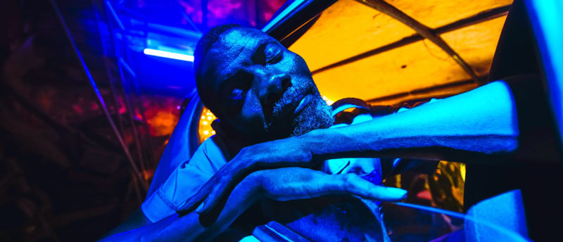 Cena do filme Ar Condicionado. Mostra de perto um homem negro de olhos fechados, cabelo curto e barba grisalha com os dois braços apoiados através da janela aberta de um carro. Ele é banhado por uma luz azul. O teto do carro é iluminado por uma luz amarela.